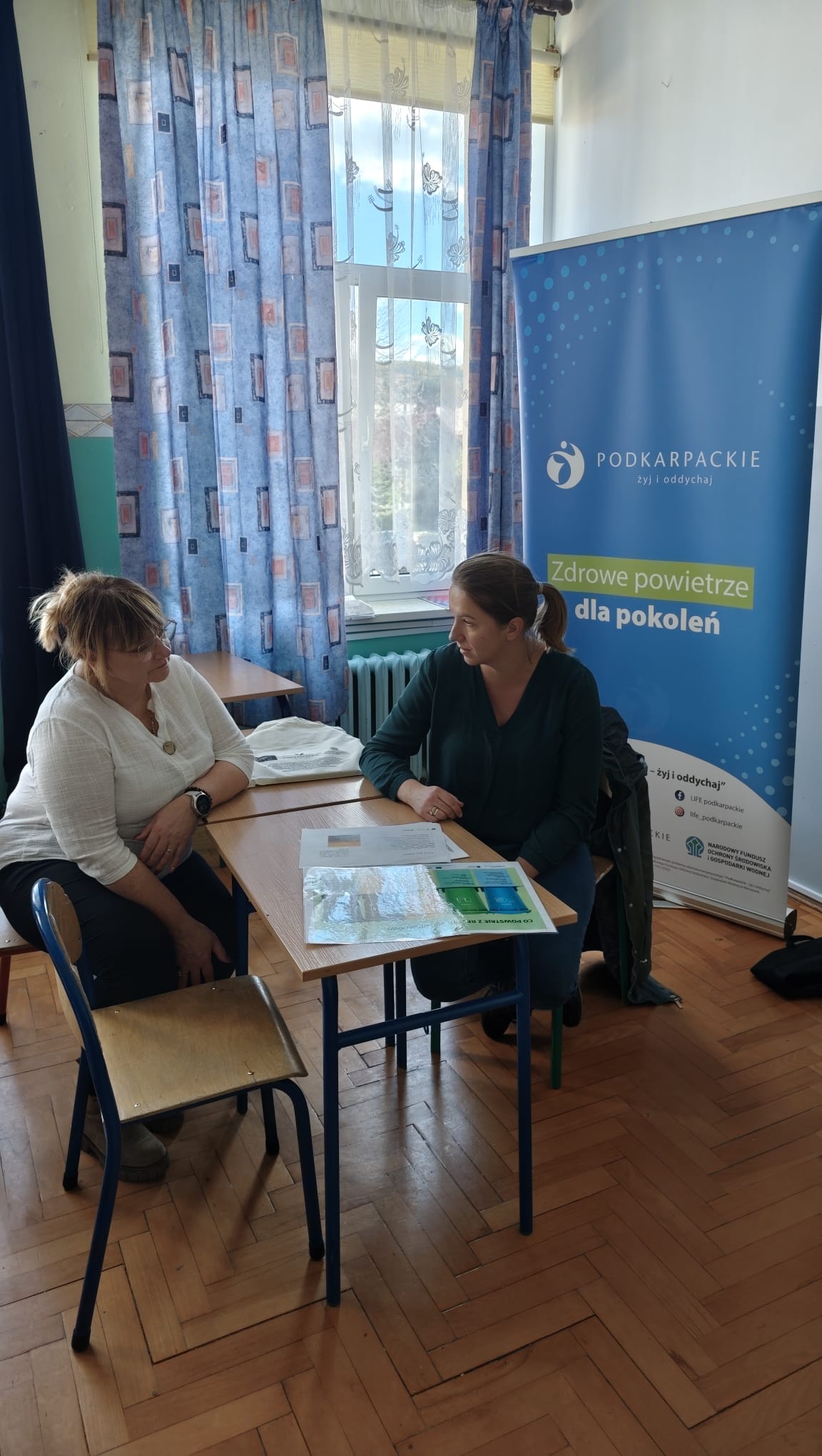 Dwie kobiety siedzą przy stole, rozmawiają. Za nimi baner z napisem "Podkarpackie. Zdrowe powietrze dla pokoleń". W tle okno z zasłonami.